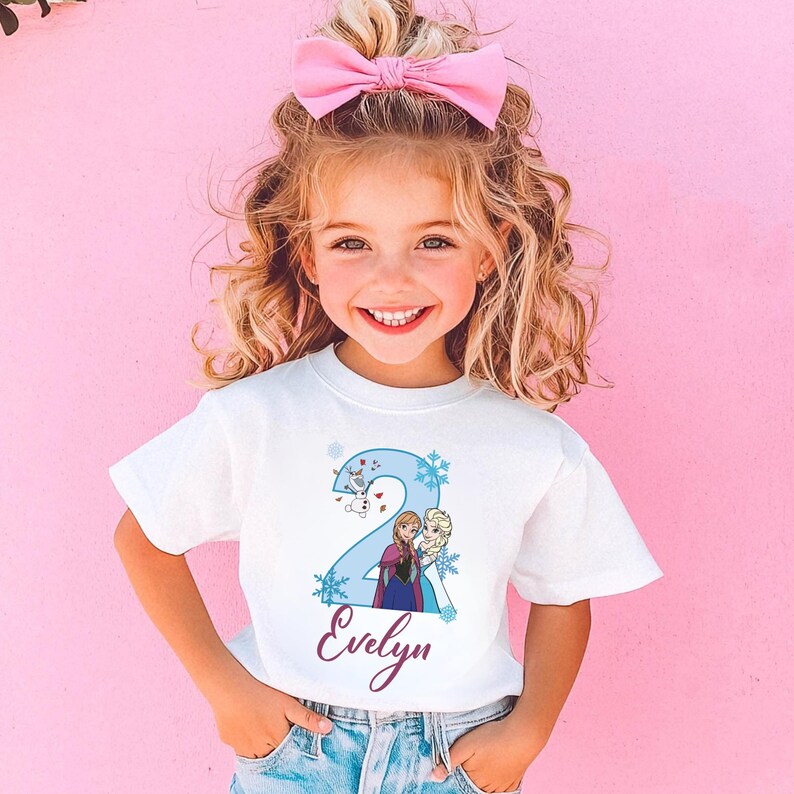 May include: A white t-shirt featuring a blue number 2 with cartoon characters and snowflakes. The name "Evelyn" is written in a cursive font below the number. The girl is wearing a pink bow.