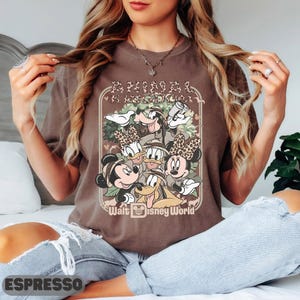May include: A brown t-shirt with a graphic of Disney characters, including Mickey and Minnie Mouse, Donald and Daisy Duck, and Goofy. The design includes the text "ANIMAL KINGDOM" and "Walt Disney World". The word "ESPRESSO" is at the bottom.