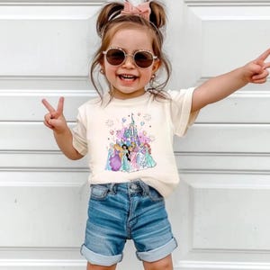 May include: A cream-colored t-shirt featuring a colorful graphic of Disney princesses in front of a castle. The shirt is paired with denim shorts. The image shows a young child wearing sunglasses and making a peace sign.