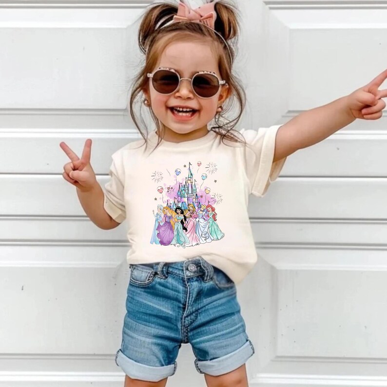 May include: A cream-colored t-shirt featuring a colorful graphic of Disney princesses in front of a castle. The shirt is paired with denim shorts. The image shows a young child wearing sunglasses and making a peace sign.