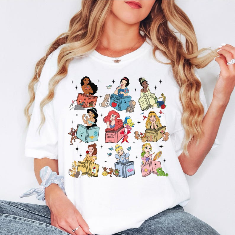Peut inclure: T-shirt blanc avec des illustrations color&eacute;es de princesses Disney lisant des livres. Chaque princesse est repr&eacute;sent&eacute;e avec son nom sur le livre, entour&eacute;e d'images associ&eacute;es. Le design comprend des personnages comme Ariel, Belle et Cendrillon.