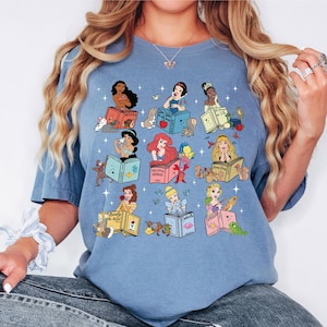 Peut inclure: T-shirt bleu clair avec des illustrations de princesses Disney lisant des livres. Chaque princesse est repr&eacute;sent&eacute;e avec son nom sur le livre, entour&eacute;e de personnages et d'objets connexes. Le t-shirt a un col rond et des manches courtes.
