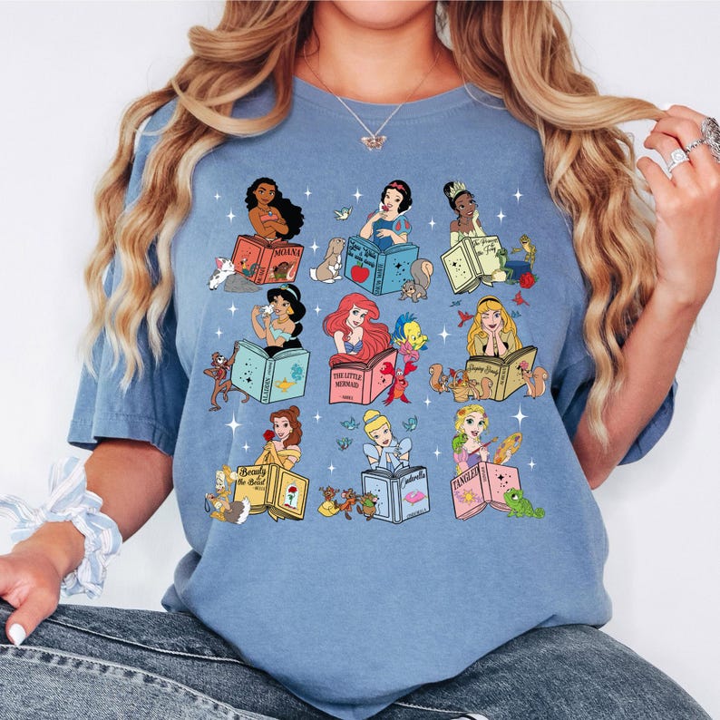 Peut inclure: T-shirt bleu clair avec des illustrations de princesses Disney lisant des livres. Chaque princesse est repr&eacute;sent&eacute;e avec son nom sur le livre, entour&eacute;e de personnages et d'objets connexes. Le t-shirt a un col rond et des manches courtes.