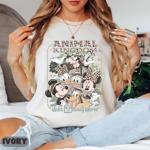 May include: Ivory t-shirt with a graphic of Disney characters, including Mickey and Minnie Mouse, Donald and Daisy Duck, Goofy, and Pluto. The text "ANIMAL KINGDOM" and "Walt Disney World" are printed on the shirt.