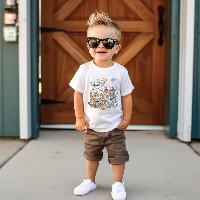 K&ouml;nnte beinhalten: Ein Kind tr&auml;gt ein wei&szlig;es T-Shirt mit einer bunten Cartoon-Grafik und dem Wort "Disney". Das Kind tr&auml;gt auch braune Shorts, wei&szlig;e Turnschuhe und eine schwarze Sonnenbrille. Der Hintergrund zeigt eine Holzt&uuml;r.