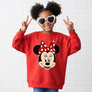May include: A red sweatshirt with a Minnie Mouse design. The graphic shows Minnie's face, a red bow with white spots, and black ears. The sweatshirt is worn by a person wearing heart-shaped sunglasses.