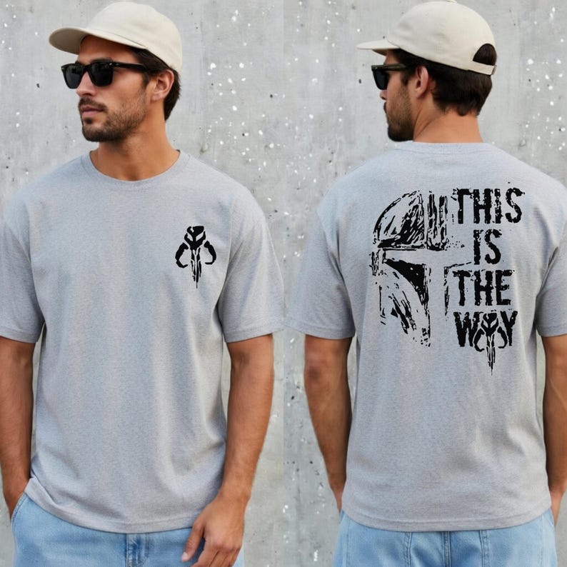 May include: A light gray short-sleeved t-shirt featuring a small black Mandalorian helmet graphic on the front left chest and a large black Mandalorian helmet graphic on the back with the text THIS IS THE WAY printed in a distressed black font.