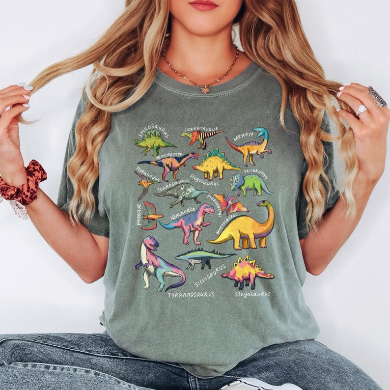 May include: A sage green t-shirt featuring a colorful dinosaur graphic. The design includes various dinosaur illustrations with their names, such as Tyrannosaurus and Stegosaurus. The shirt is a relaxed fit.