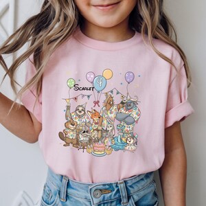 K&ouml;nnte beinhalten: Ein hellrosa T-Shirt mit einer Geburtstagsparty-Szene mit Zeichentierfiguren. Das Shirt hat den Namen "Scarlet" und die Zahl "4" sowie Luftballons und einen Kuchen. Das Shirt wird von einem Kind getragen.