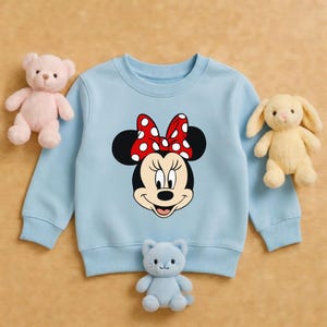 May include: A pale blue sweatshirt with a Minnie Mouse design, showcasing a red bow with white spots. The image includes a pink teddy bear, a yellow bunny, and a blue cat soft toy.