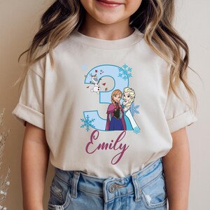 May include: Beige t-shirt featuring a blue number 3 with snowflakes and cartoon characters Anna, Elsa, and Olaf. The name "Emily" is written in a cursive font below the number. The shirt is designed for a child's third birthday.