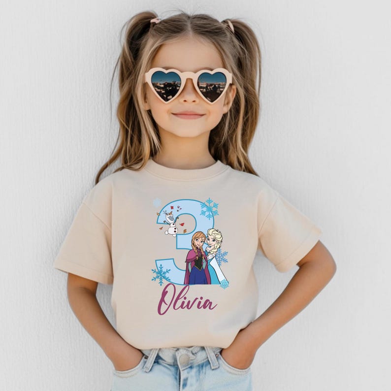May include: A beige t-shirt featuring a blue number 3 with snowflakes and cartoon characters from the movie Frozen. The name "Olivia" is written in a cursive font below the number. The child is wearing heart-shaped sunglasses.