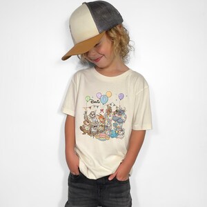 K&ouml;nnte beinhalten: Ein cremefarbenes T-Shirt mit einer farbenfrohen Illustration von Zeichentieren, die einen Geburtstag feiern. Das Shirt tr&auml;gt den Namen "Davis" &uuml;ber der Illustration. Die Person tr&auml;gt eine grau-braune Baseballkappe.