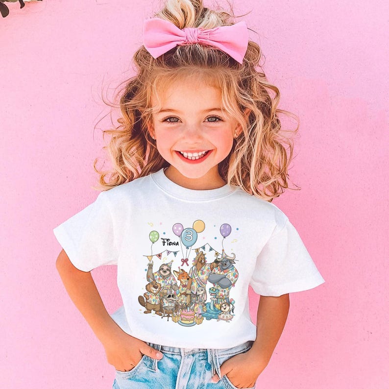 K&ouml;nnte beinhalten: Ein wei&szlig;es T-Shirt mit einer farbenfrohen Illustration von Zeichentieren, die einen Geburtstag feiern. Auf dem Shirt stehen der Name "Fiona" und die Zahl "3". Das M&auml;dchen tr&auml;gt eine rosa Schleife im Haar und l&auml;chelt.