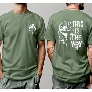 May include: An olive green short-sleeve t-shirt featuring a white Mandalorian helmet icon on the left chest. The back of the shirt displays a large white graphic of a Mandalorian helmet with the text THIS IS THE WAY printed in a distressed font.