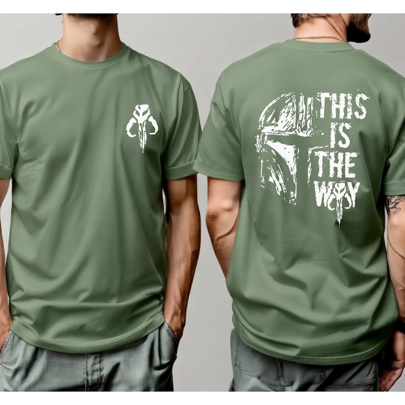 May include: An olive green short-sleeve t-shirt featuring a white Mandalorian helmet icon on the left chest. The back of the shirt displays a large white graphic of a Mandalorian helmet with the text THIS IS THE WAY printed in a distressed font.