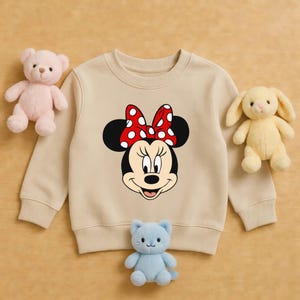 May include: A beige sweatshirt with a Minnie Mouse design, featuring a red bow with white spots. The sweatshirt is surrounded by a pink teddy bear, a yellow bunny, and a blue cat soft toy.