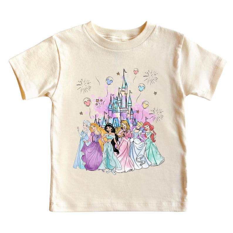 May include: Cream-colored short-sleeved t-shirt featuring a colorful graphic of several princesses in front of a castle. The graphic includes fireworks and balloon-like shapes. The princesses are wearing gowns in various colors.