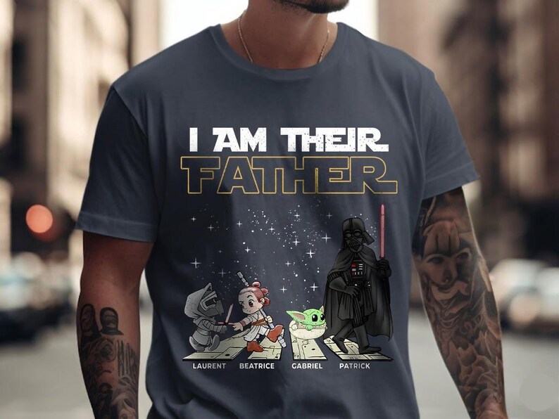 May include: Navy blue t-shirt with the text "I AM THEIR FATHER" in white and gold. The shirt features cartoon illustrations of Star Wars characters, including Darth Vader, and the names Laurent, Beatrice, Gabriel, and Patrick.