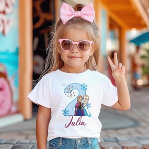 May include: A white t-shirt featuring a blue number 2 design with snowflakes, and cartoon characters. The name "Julia" is written in a purple script font. The girl is wearing pink sunglasses and a pink bow.