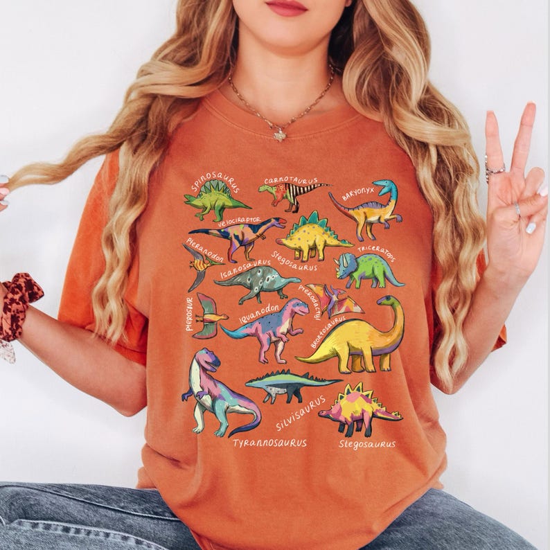 May include: Orange t-shirt featuring colorful dinosaur illustrations with their names. The dinosaurs include Stegosaurus, Tyrannosaurus, and Triceratops. The shirt is a comfortable, casual style.