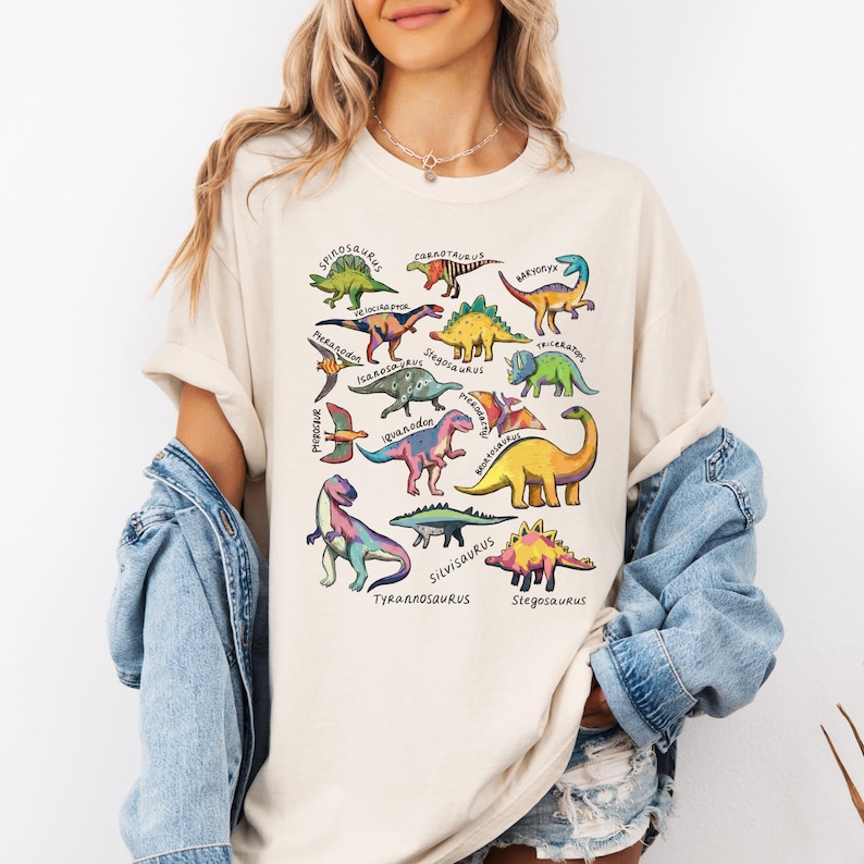 May include: Cream-colored t-shirt featuring colorful dinosaur illustrations with their names. The dinosaurs are in various colors, including green, yellow, blue, and pink. The shirt has a relaxed fit and short sleeves, perfect for casual wear.
