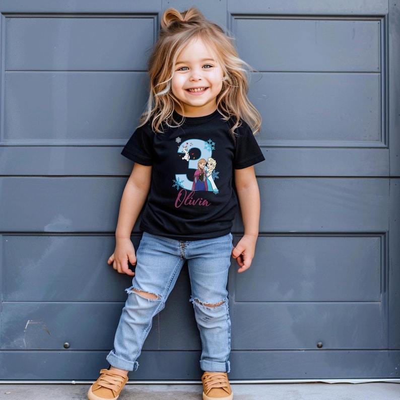 May include: A young child wearing a black t-shirt with a large number 3 and the name "Olivia" printed on it. The shirt features a graphic of two animated characters. The child is also wearing blue jeans and brown shoes.