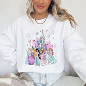 May include: White sweatshirt featuring a colorful illustration of a castle and several princesses. The design includes pink, blue, and purple accents, with fireworks and balloons. The princesses are wearing gowns in various pastel colors.