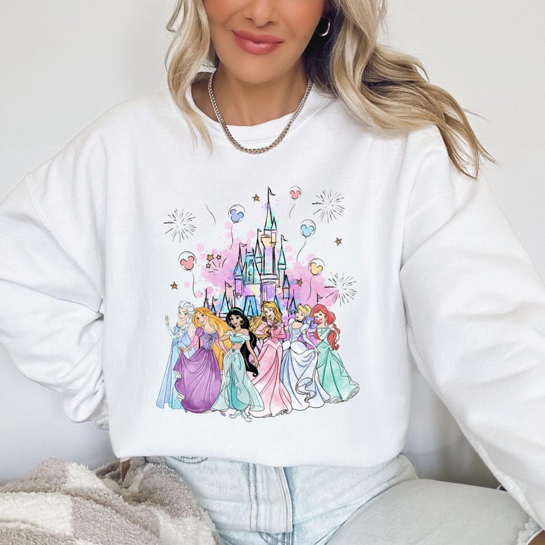 May include: White sweatshirt featuring a colorful illustration of a castle and several princesses. The design includes pink, blue, and purple accents, with fireworks and balloons. The princesses are wearing gowns in various pastel colors.