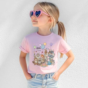 K&ouml;nnte beinhalten: Ein hellrosa T-Shirt mit einer Geburtstagsparty-Grafik mit Tierfiguren, Luftballons und einer Torte. Der Name "Olivia" ist auf dem T-Shirt aufgedruckt. Das M&auml;dchen tr&auml;gt eine herzf&ouml;rmige Sonnenbrille in Rosa und eine blaue Jeans.