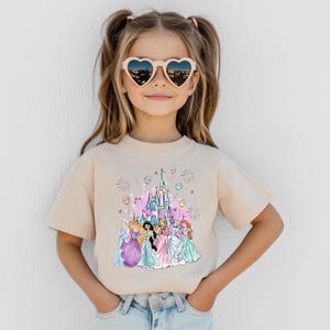 May include: A beige t-shirt featuring a colorful graphic of a castle and several princesses. The child is wearing heart-shaped sunglasses. The graphic includes pink, blue, and purple elements, with fireworks and balloons.