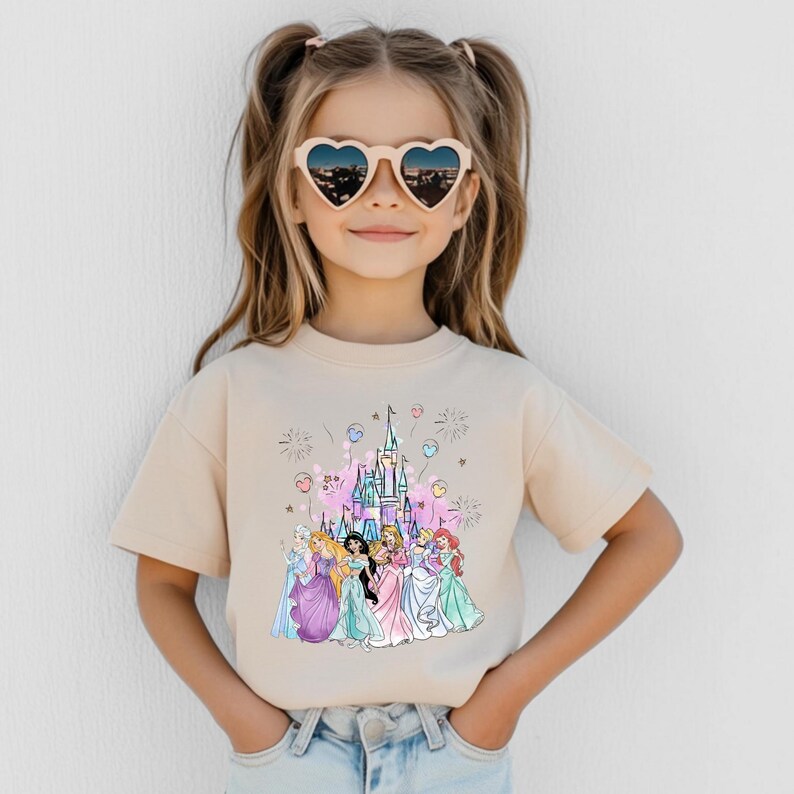 May include: A beige t-shirt featuring a colorful graphic of a castle and several princesses. The child is wearing heart-shaped sunglasses. The graphic includes pink, blue, and purple elements, with fireworks and balloons.