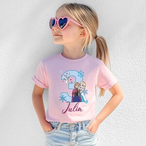 May include: A pink t-shirt featuring a blue number 3 with snowflakes and cartoon characters. The name "Julia" is written in cursive below the number. The shirt is worn by a child wearing pink heart-shaped sunglasses.