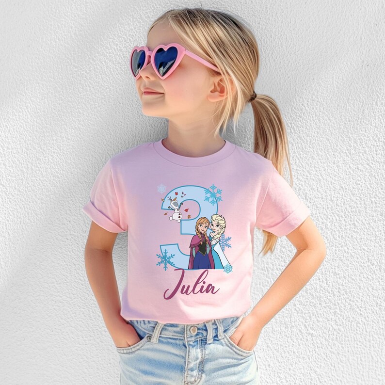 May include: A pink t-shirt featuring a blue number 3 with snowflakes and cartoon characters. The name "Julia" is written in cursive below the number. The shirt is worn by a child wearing pink heart-shaped sunglasses.