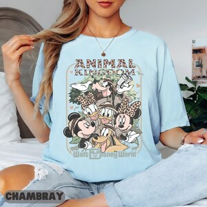 May include: Pale blue t-shirt with a graphic of Disney characters, including Mickey and Minnie Mouse, Donald Duck, and Goofy. The shirt has the text "ANIMAL KINGDOM" and "Walt Disney World". The shirt is made of a soft material.