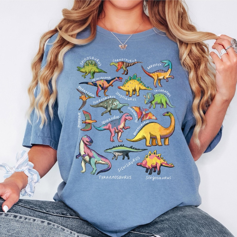 May include: A light blue t-shirt featuring colorful dinosaur illustrations with their names. The dinosaurs are in various colors, including green, yellow, pink, and blue. The shirt has a relaxed fit and short sleeves.