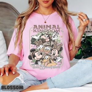 May include: A pale pink t-shirt with a graphic of Disney characters, including Mickey and Minnie Mouse, Donald Duck, Goofy, and Pluto. The text "ANIMAL KINGDOM" and "Walt Disney World" are also displayed.