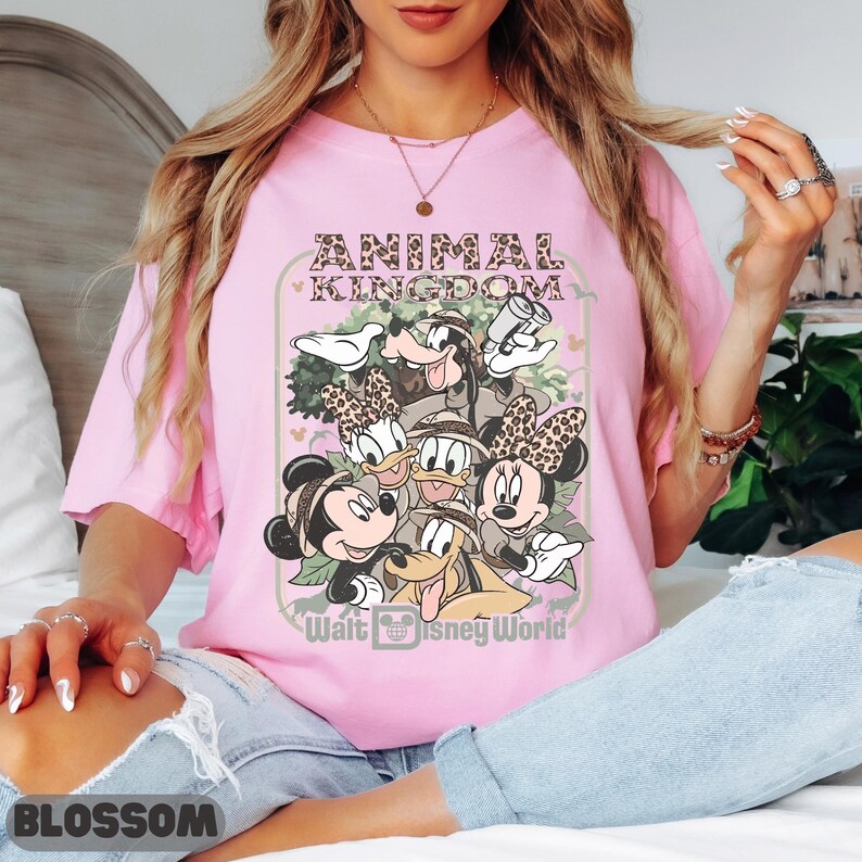 May include: A pale pink t-shirt with a graphic of Disney characters, including Mickey and Minnie Mouse, Donald Duck, Goofy, and Pluto. The text "ANIMAL KINGDOM" and "Walt Disney World" are also displayed.