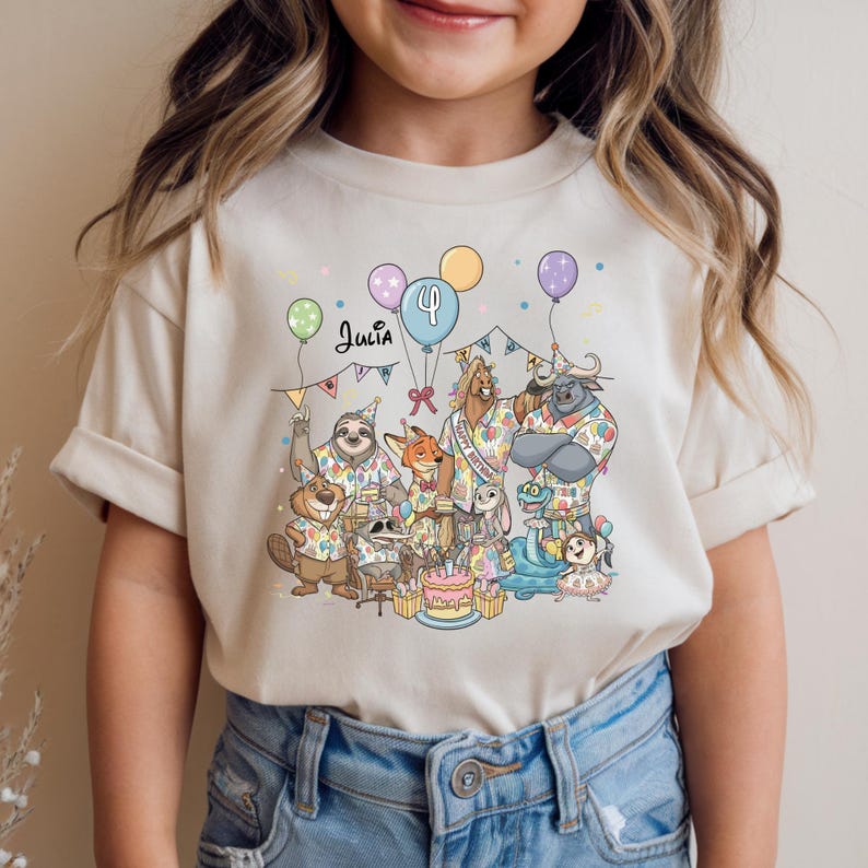 K&ouml;nnte beinhalten: Beiges T-Shirt mit einer bunten Geburtstagsparty-Szene mit Zeichentierfiguren, Luftballons und einer Torte. Der Name "Julia" steht &uuml;ber den Tieren, mit der Zahl "4" auf einem Ballon.
