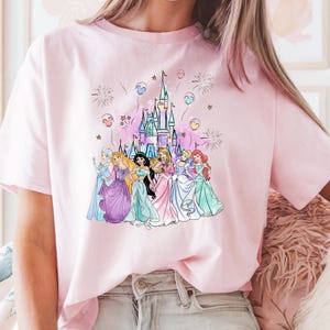 May include: A light pink t-shirt featuring a colorful graphic of a fairytale castle, fireworks, and several princesses. The princesses are wearing gowns in various colors. The shirt is made of a soft material.