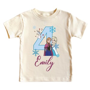 May include: A cream-colored t-shirt featuring a blue number 4 with illustrations of Anna, Elsa, and Olaf from Frozen. The name "Emily" is written in cursive below the number. The shirt is designed for a child.