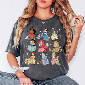 Peut inclure: T-shirt gris fonc&eacute; avec des illustrations de princesses Disney lisant des livres. Chaque princesse est repr&eacute;sent&eacute;e avec ses compagnons animaux et les titres des livres. Le design comprend des &eacute;toiles et une esth&eacute;tique fantaisiste.