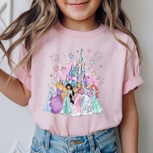 May include: Pink t-shirt featuring a colorful illustration of a castle and several princesses. The princesses are wearing gowns in various colors, and the design includes fireworks and heart-shaped balloons.