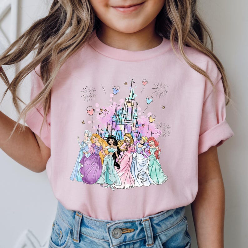 May include: Pink t-shirt featuring a colorful illustration of a castle and several princesses. The princesses are wearing gowns in various colors, and the design includes fireworks and heart-shaped balloons.