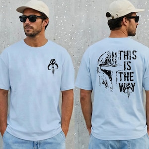 May include: A light blue t-shirt featuring a small black Mandalorian helmet graphic on the front left chest. The back of the shirt displays a large black Mandalorian helmet graphic with the text "THIS IS THE WAY" in a distressed font.