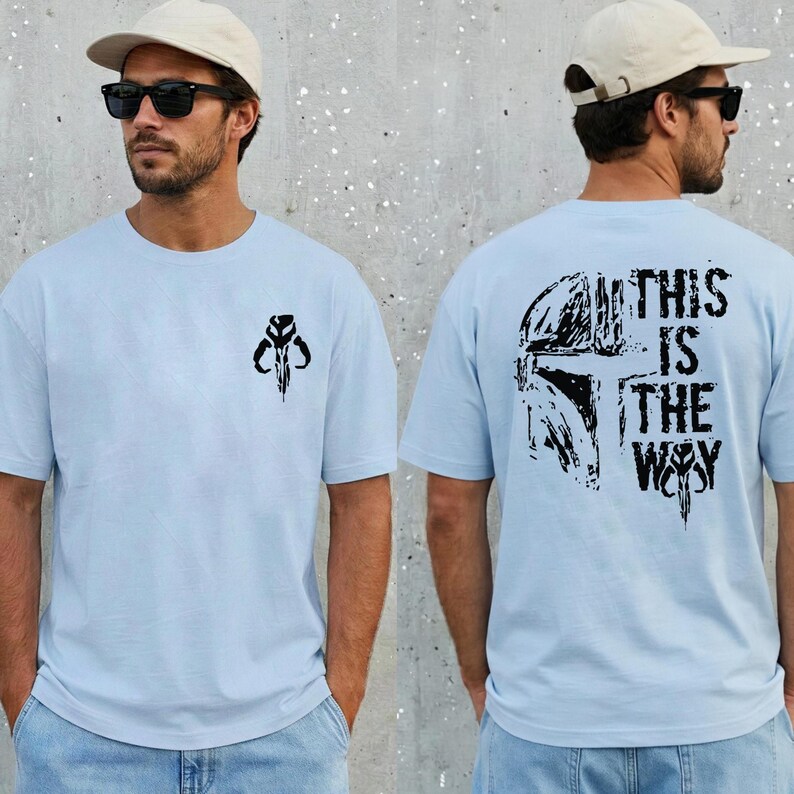 May include: A light blue t-shirt featuring a small black Mandalorian helmet graphic on the front left chest. The back of the shirt displays a large black Mandalorian helmet graphic with the text "THIS IS THE WAY" in a distressed font.