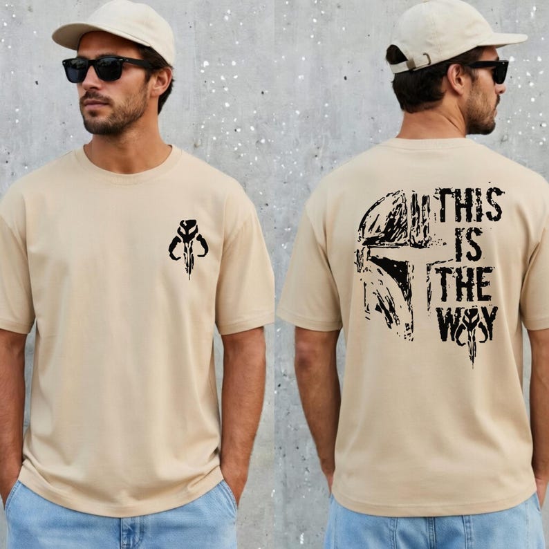 May include: A beige short-sleeve t-shirt featuring a small black Mandalorian helmet icon on the left chest. The back of the shirt displays a large black graphic of a Mandalorian helmet with the text THIS IS THE WAY in a distressed font.