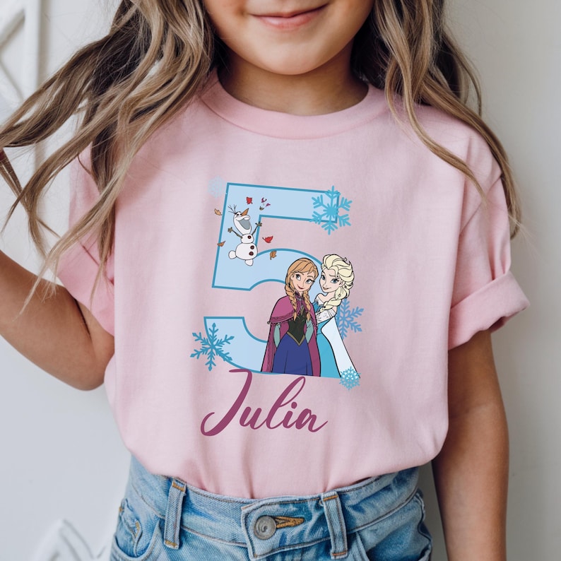 May include: Pink t-shirt featuring a blue number 5 with snowflakes and cartoon images of Anna, Elsa, and Olaf. The name "Julia" is written in cursive below the number. The shirt is worn by a child.