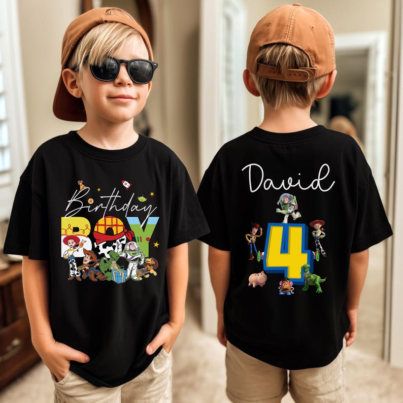Pode incluir: T-shirt preto de manga curta com um gr&aacute;fico colorido "Birthday Boy" com personagens de desenhos animados. A parte de tr&aacute;s da camisa tem o nome "David" e o n&uacute;mero "4" com mais personagens de desenhos animados. O menino est&aacute; usando um bon&eacute; de beisebol marrom e &oacute;culos de sol.