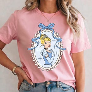 K&ouml;nnte beinhalten: Hellrosa T-Shirt mit einem Cinderella-Motiv. Das Motiv ist ein ovales Bild mit hellblauem Hintergrund, einem Portr&auml;t von Cinderella und einer blauen Schleife. Die Prinzessin tr&auml;gt eine Tiara und ein blaues Kleid.